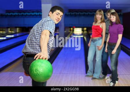 Young man prepares throw ball on path for bowling and three girls look on him, focus on man Stock Photo