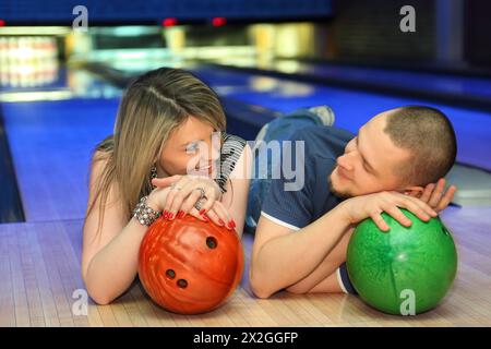 Fellow and girl lie on parquet and look on each other leaning against balls for bowling, focus on girl Stock Photo