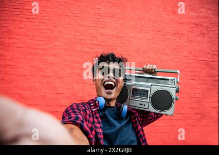 Smiling young man with boombox celebrates life. Vibrant street culture shines against a red wall backdrop. Energy and retro vibes in urban setting. Stock Photo
