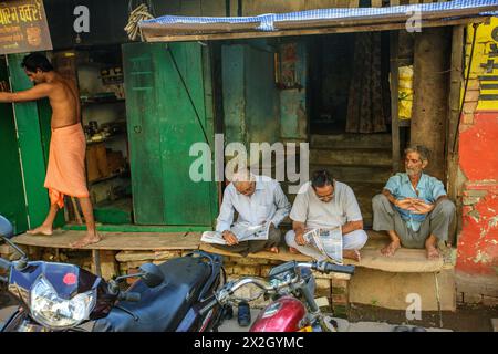 Men reading newspapers as a man wearing a dhoti opens the shutters o his shop in Varanasi, India Stock Photo