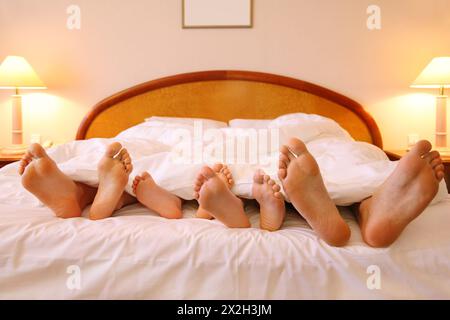 Mother, father and two children lie on bed with white sheets; focus on feet Stock Photo