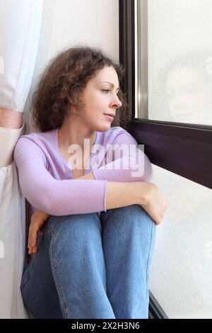 Young beutiful woman sits on windowsill, looks out window and dreams Stock Photo
