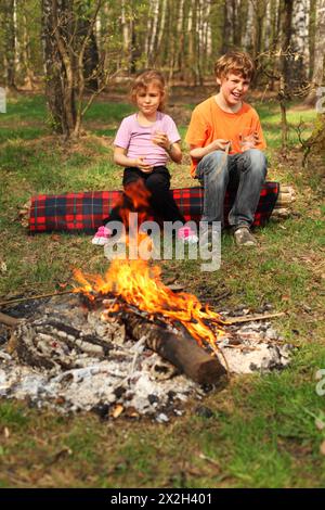 Two children sit near campfire and hold in their hands glasses; focus on boy Stock Photo