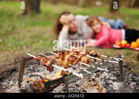 Mother and daughter lie near campfire with grill and barbecue; focus on meat Stock Photo