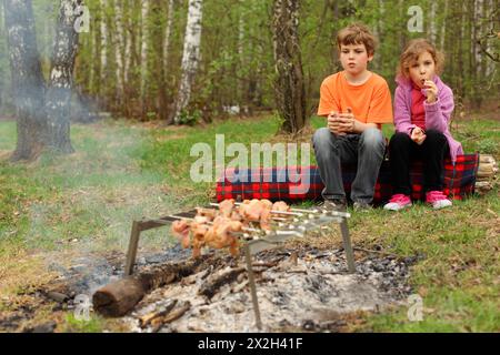 Two children sit near campfire with grill and barbecue; brother holds glass, sister sucks lollipop Stock Photo