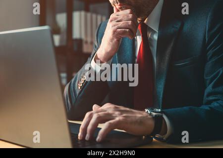 Worried businessman working on laptop computer late at night in dark office, thinking with hand on the chin, selective focus Stock Photo