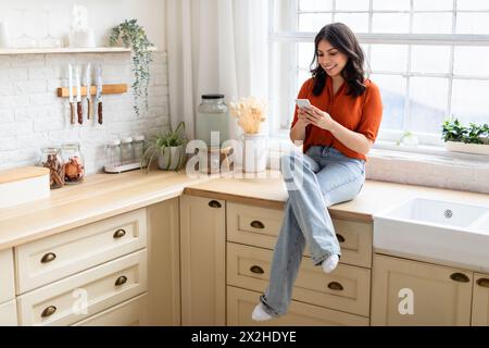 Woman sitting on kitchen counter using smartphone Stock Photo