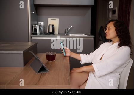 Relaxed young multi ethnic woman in white bathrobe, using mobile phone at home, checking social media content, new mobile application, booking, orderi Stock Photo