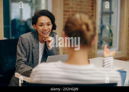 Two smiling female freelancers talking during work on laptop while sitting in cozy coworking Stock Photo