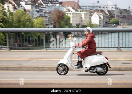 Basel, Switzerland - April 18, 2024: One woman wearing parka riding white scooter motorbike on Wettsteinbrucke Stock Photo