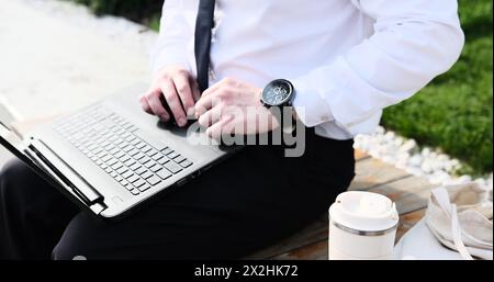 Caucasian bearded business man freelancer working on break with laptop outdoors in park. Male office worker sitting on a bench on a city street park Stock Photo