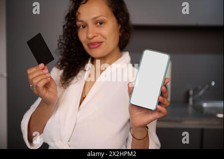 Young woman holds and shows at camera blank black credit card and smartphone with white empty mockup digital screen, makes online payments, transferri Stock Photo