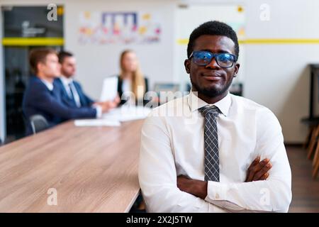 Confident African corporate professional in formal workwear sits at meeting table, focus on leadership with diverse team blurred in background Stock Photo