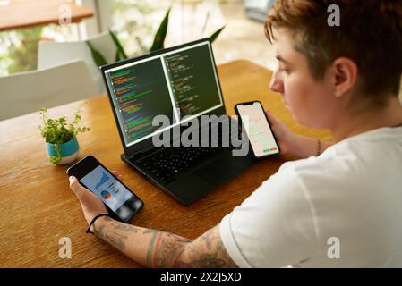 Person at wooden table multitasks with coding on laptop, analyzes data on two smartphones. Concentrated individual with tattoos uses tech for Stock Photo