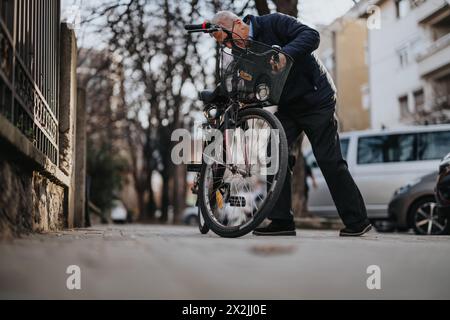 Elderly man attentively fixing his bike on a city sidewalk, depicting active senior lifestyle and urban commuting. Stock Photo