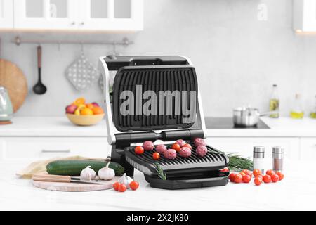 Electric grill with meatballs, tomatoes and vegetables on white marble table in kitchen Stock Photo