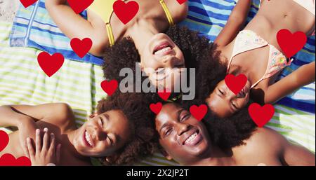 Image of red hearts over smiling african american family on holiday lying on beach towels Stock Photo