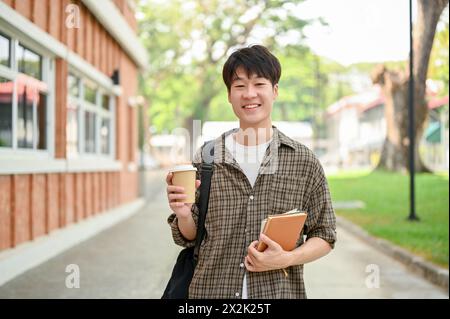 A smiling, positive young Asian man college student, carrying a backpack, stands in front of a brick building on campus, holding a coffee cup and a bo Stock Photo