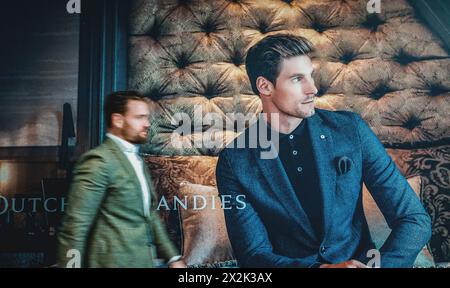 A sharply dressed man in a refined blue blazer stands confidently at an opulent indoor setting, exuding style and sophistication while another man wal Stock Photo