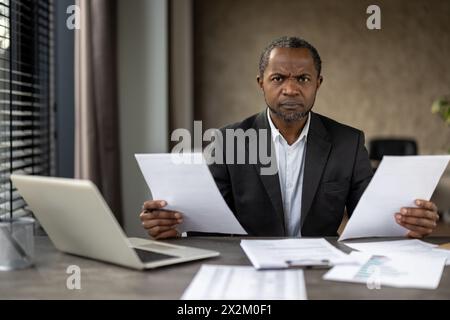 Mature African American businessman in a suit frowns as he reviews important documents, reflecting a stressful moment in a professional setting. Stock Photo