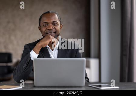 A professional black businessman smiles confidently in a modern office setting, using a laptop at his desk surrounded by technology. Stock Photo