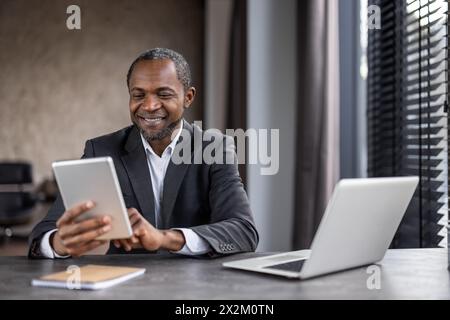 A cheerful professional black businessman in a suit sits at a modern office desk, happily using his tablet with a laptop nearby. Stock Photo