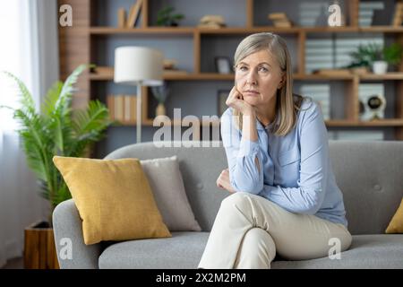 A thoughtful looking mature woman sitting in her cozy living room, gazing into the distance. The peaceful home environment reflects a moment of solitude or contemplation. Stock Photo