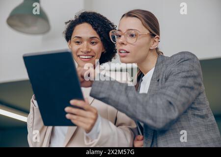 Smiling female coworkers discussing project standing in modern office and use digital tablet Stock Photo