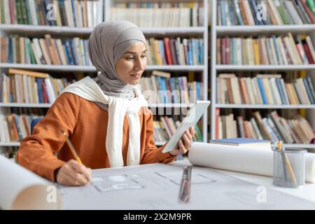 A young female architect wears a hijab while focusing on her project designs using a digital tablet in a library setting, surrounded by books. Stock Photo