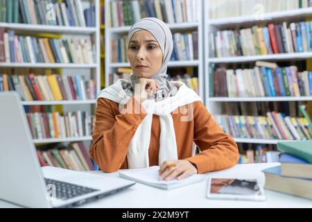 A thoughtful Muslim woman in a hijab engages with her studies using a laptop in a public library, surrounded by books. Stock Photo