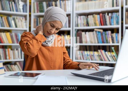 A young Muslim woman experiencing neck pain as she works at her laptop in a library setting. The image captures a moment of discomfort amidst a study session. Stock Photo