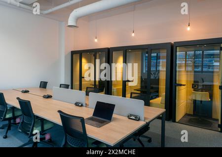 A contemporary office space featuring a long meeting table with laptops and a row of private glass booths with yellow chairs Stock Photo