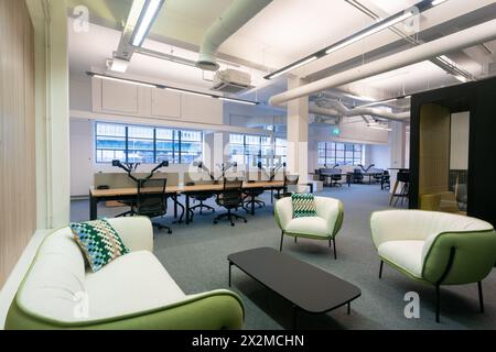A spacious and brightly lit modern office featuring an open-plan workspace with ergonomic chairs and large tables, complemented by cozy lounge areas w Stock Photo