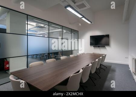 A contemporary office conference room featuring a long table with chairs, glass walls, and a wall-mounted monitor for presentations Stock Photo
