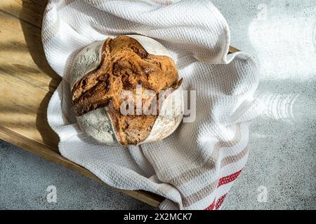 A freshly baked sourdough loaf made with rye flour, resting on a wooden board covered with a white kitchen towel Stock Photo