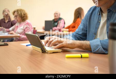 Elderly students engaged in an educational setting, focusing on a man using a tablet with peers in the background Stock Photo