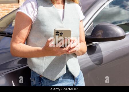 A woman engages with her phone, standing outside next to her vehicle, on a sunny day Stock Photo
