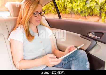 A professional woman in glasses focused on her tablet while sitting in the backseat of a car, with vineyards outside Stock Photo