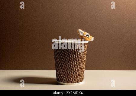 A creative composition showing a small toy fawn peeking from a textured takeaway coffee cup against a beige backdrop. Stock Photo