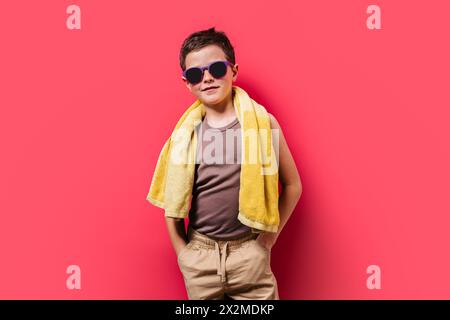 A confident young boy poses with hands on hips, wearing sunglasses and a towel over his shoulders against a vibrant pink backdrop Stock Photo