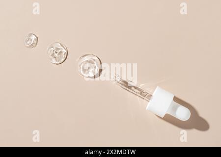 Overhead view of translucent cosmetic gel drops alongside a glass pipette on a neutral beige surface, showcasing skincare product textures Stock Photo