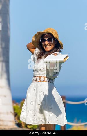 A cheerful woman wearing a straw hat and sunglasses is holding a book, smiling broadly in a natural outdoor setting. Stock Photo