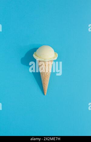 A vibrant Cream ice cream scoop sits atop a classic waffle cone against a blue background, casting a playful shadow Stock Photo