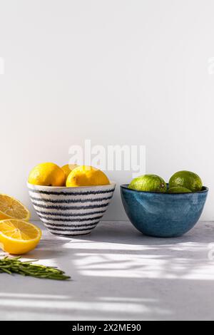 Two ceramic bowls, one with bright yellow lemons and the other with green limes, are set on a light table with soft shadows Stock Photo