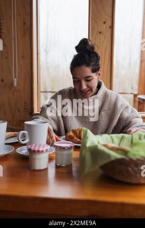 Cozy morning scene with a smiling woman enjoying breakfast at a wooden table near a window Stock Photo