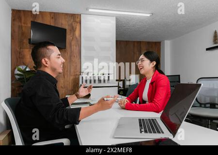 A positive work interaction between a Latin American male and female in a contemporary office setting, sharing ideas over laptops. Stock Photo