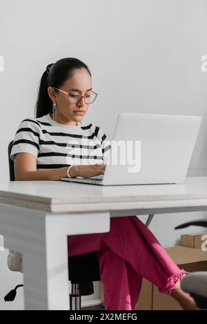 A focused Latin American female in a striped shirt works diligently on her laptop in an office setting, embodying professionalism and concentration. Stock Photo
