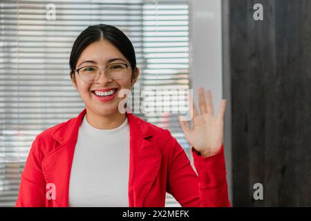 A cheerful Latin American woman wearing glasses and a red jacket, smiling and waving in a professional office setting. Stock Photo