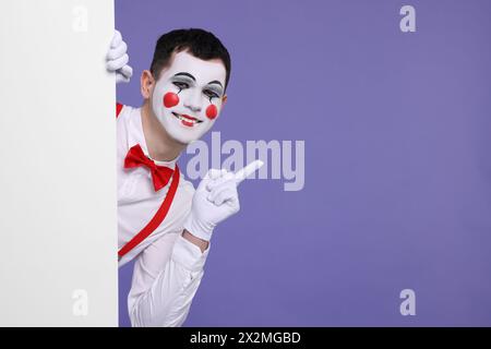 Funny mime artist peeking out of blank poster and pointing at something on purple background. Space for text Stock Photo