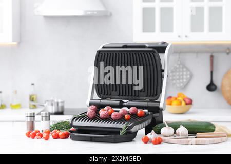 Electric grill with meatballs, tomatoes and vegetables on white marble table in kitchen Stock Photo
